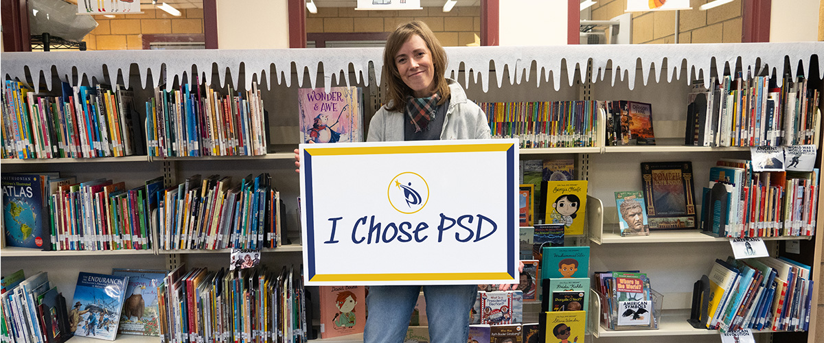 PSD alumna Annie Herzig holds an "I chose PSD" sign in a school library.