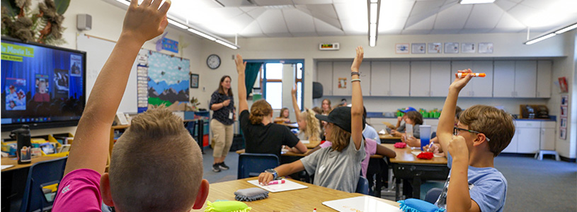 Middle school students in a classroom with hands raised.