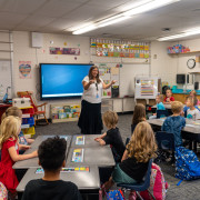 A teacher speaks at the front of an elementary classroom with a blue screen behind her